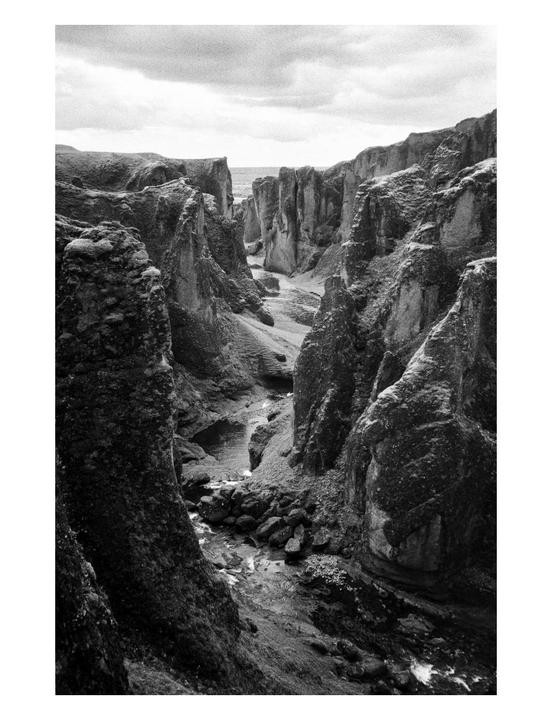 A narrow canyon with a stream running through it is surrounded by steep, rocky walls under a cloudy sky.