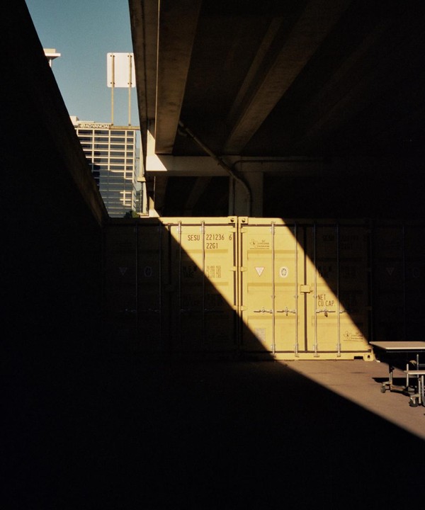 A strong diagonal beam of sunlight illuminates a yellow shipping container under a concrete overpass.