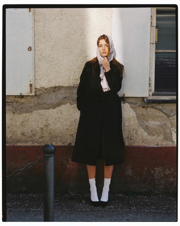 A woman wearing a headscarf and a black coat stands against a textured wall with a window and a closed shutter.