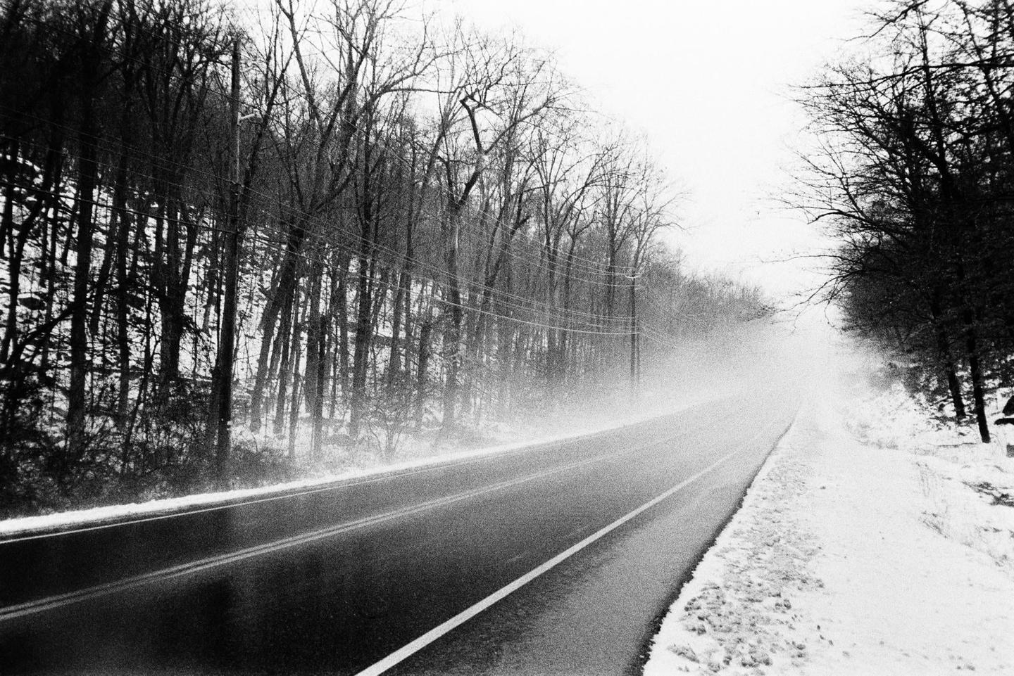 A road with snow on the side is surrounded by bare trees and telephone poles.