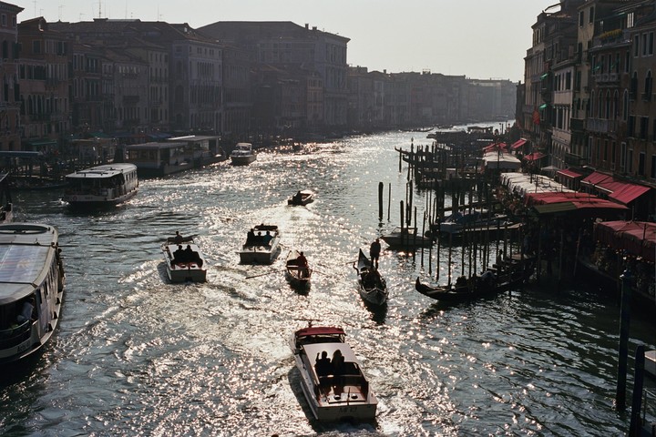 Boats and gondolas travel along a canal lined with buildings and docks under bright sunlight.