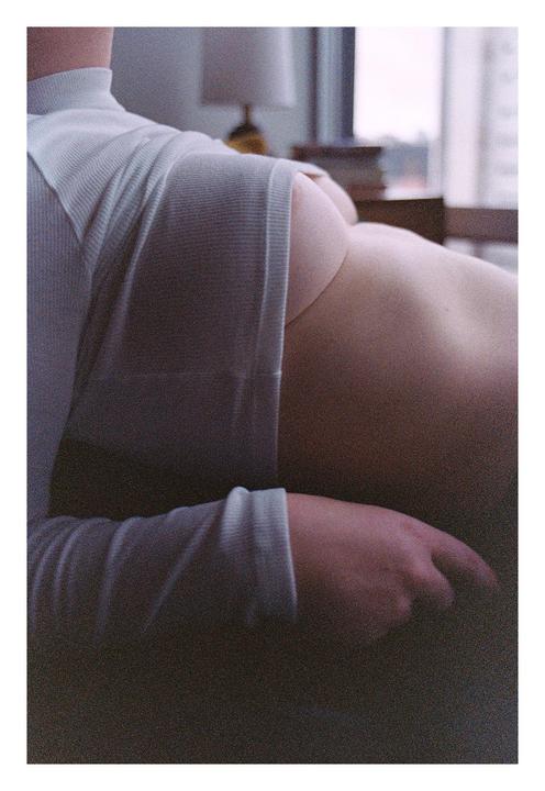 A woman is seen from the back, partially clothed in a light-colored top, with a lamp and window in the background.
