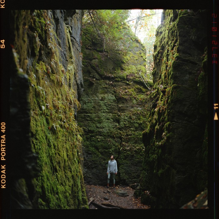 A woman stands between two moss-covered rock walls in a forest.
