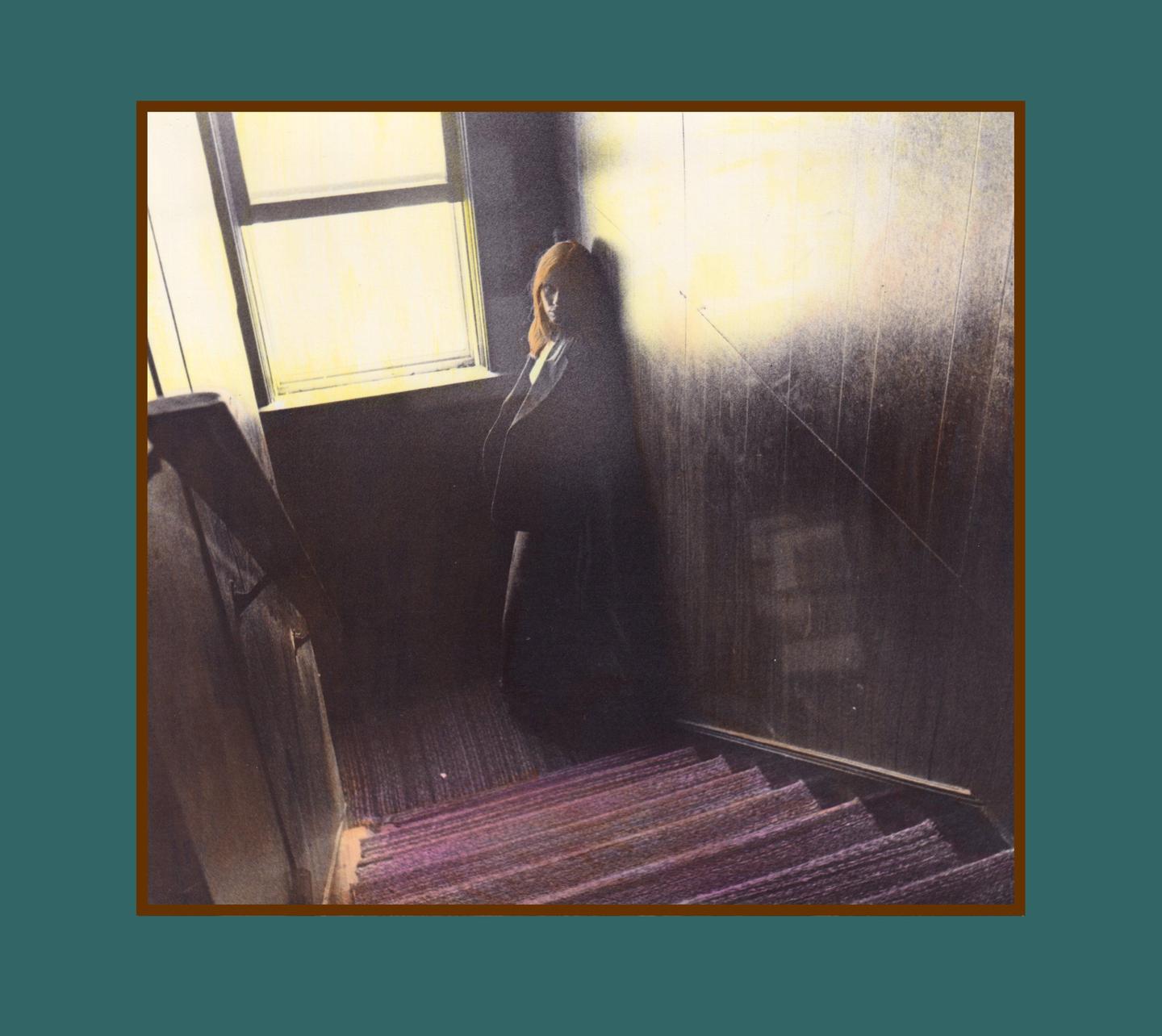 A woman stands in a dark stairwell next to a window, with light illuminating the wall behind her.
