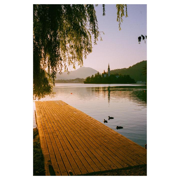 A wooden pier extends into a lake with a church on an island and mountains in the background under a golden sky.