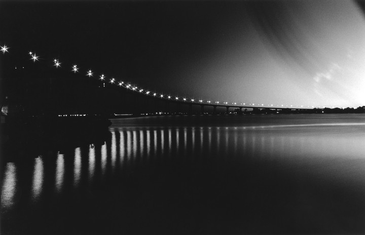 A long exposure captures a bridge illuminated by many lights over a body of water at night.