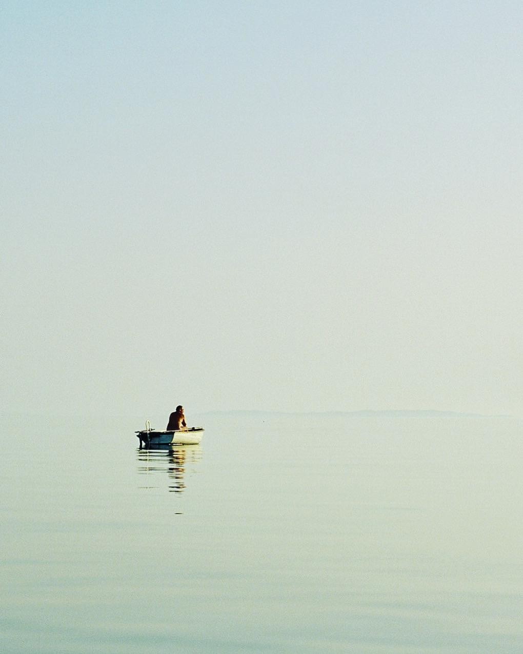 A man sits in a small boat on calm water under a clear sky.