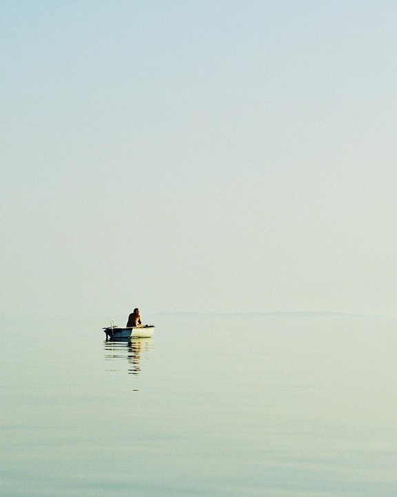 A man sits in a small boat on calm water under a clear sky.
