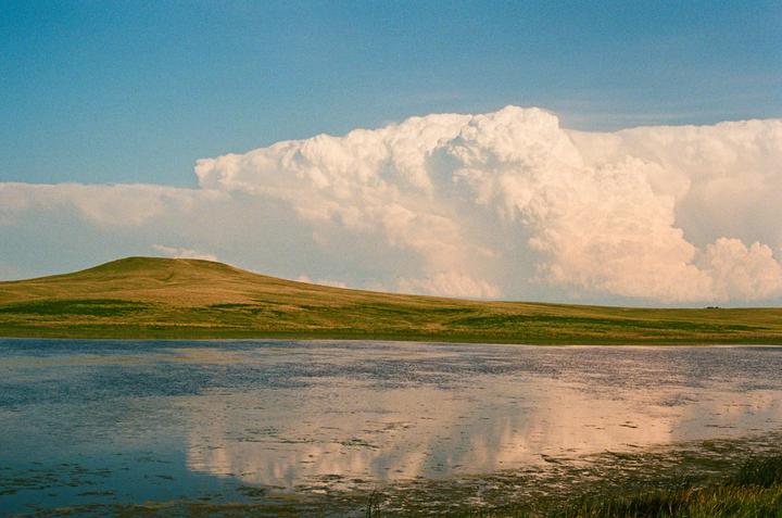 A large body of water reflects a cloudy sky and a grassy hill under a bright blue sky.