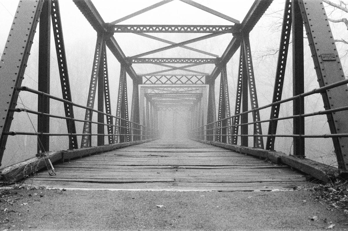 A long, narrow metal bridge with a wooden deck extends into a thick fog.