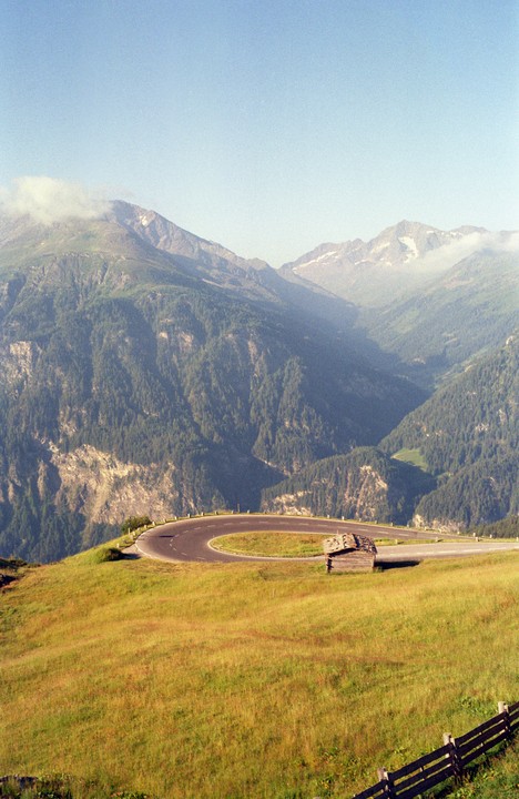 A winding road curves through a grassy mountain landscape with a small wooden hut and distant peaks.