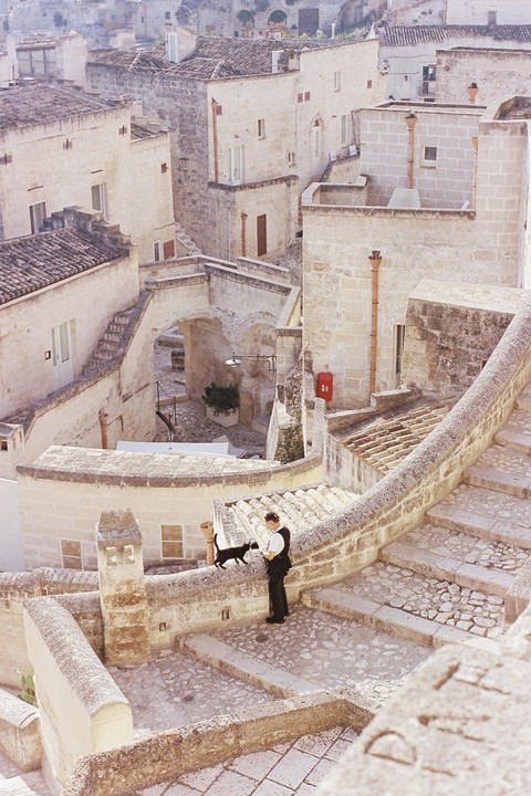 A man stands on a stone wall next to a black cat in a village of stone buildings and staircases.
