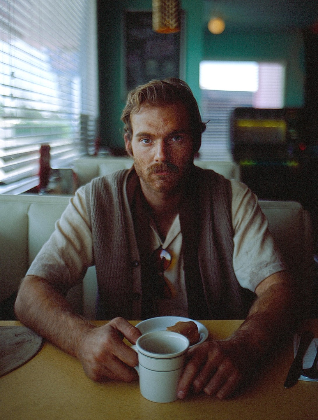 A man with a mustache sits at a diner booth with a coffee cup and a pastry.