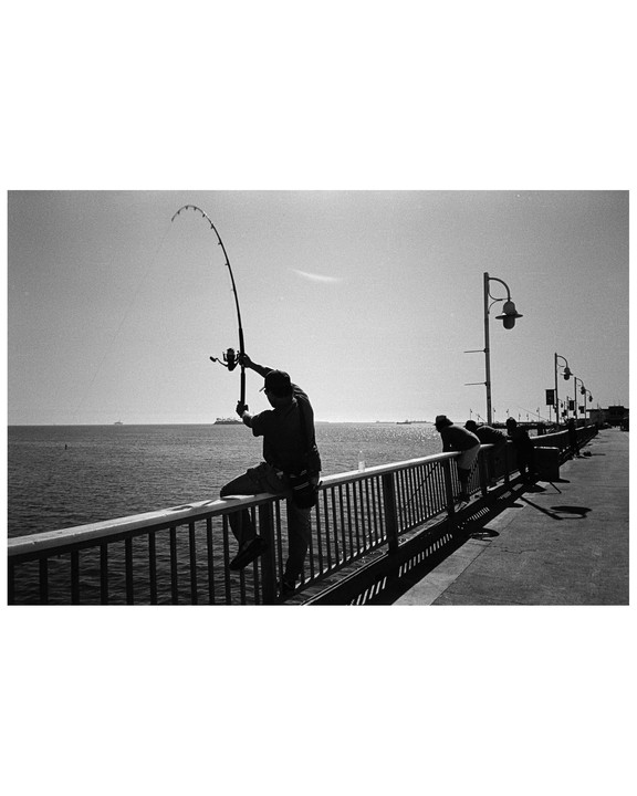 A man sits on a railing and casts a fishing rod into the water while other people fish nearby.