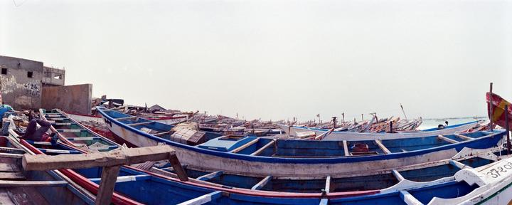 A man rests in a blue and white boat among many other boats on a beach next to buildings.