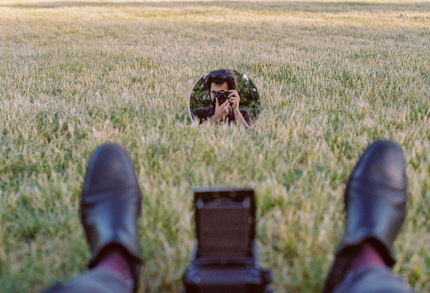 A man photographs himself in a mirror placed in a field of tall grass, with his feet visible in the foreground.