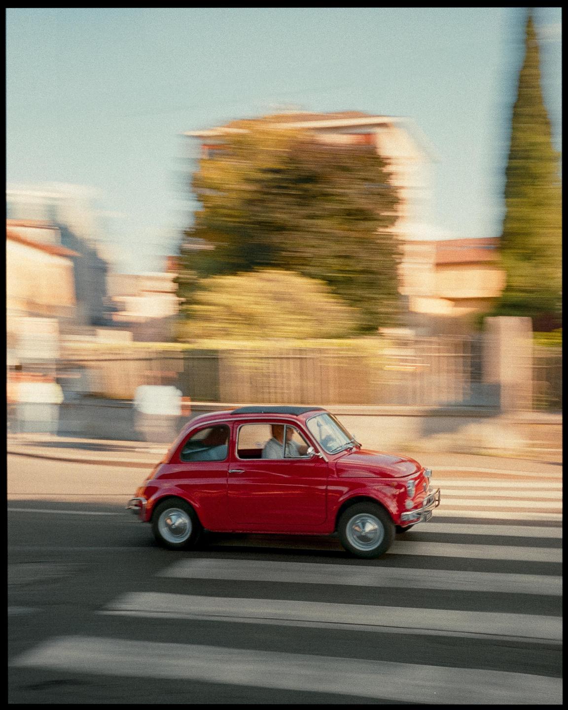 A red car drives across a crosswalk with blurred buildings and trees in the background.
