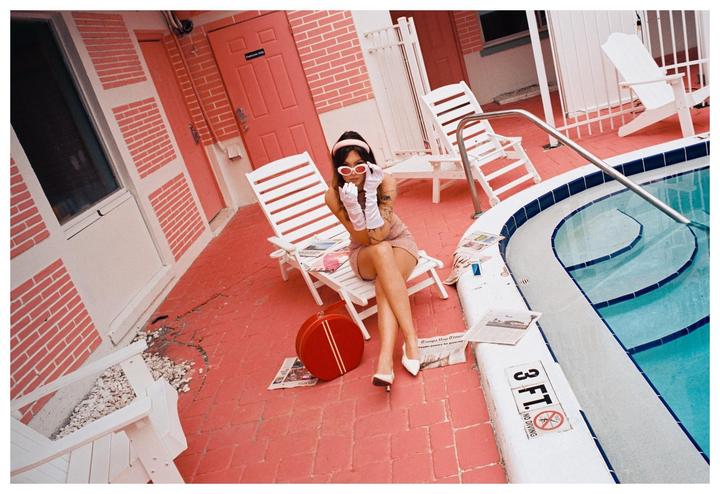 A woman in a pink dress, white gloves, and red sunglasses sits on a lounge chair next to a swimming pool with pink brick walls and pavement.