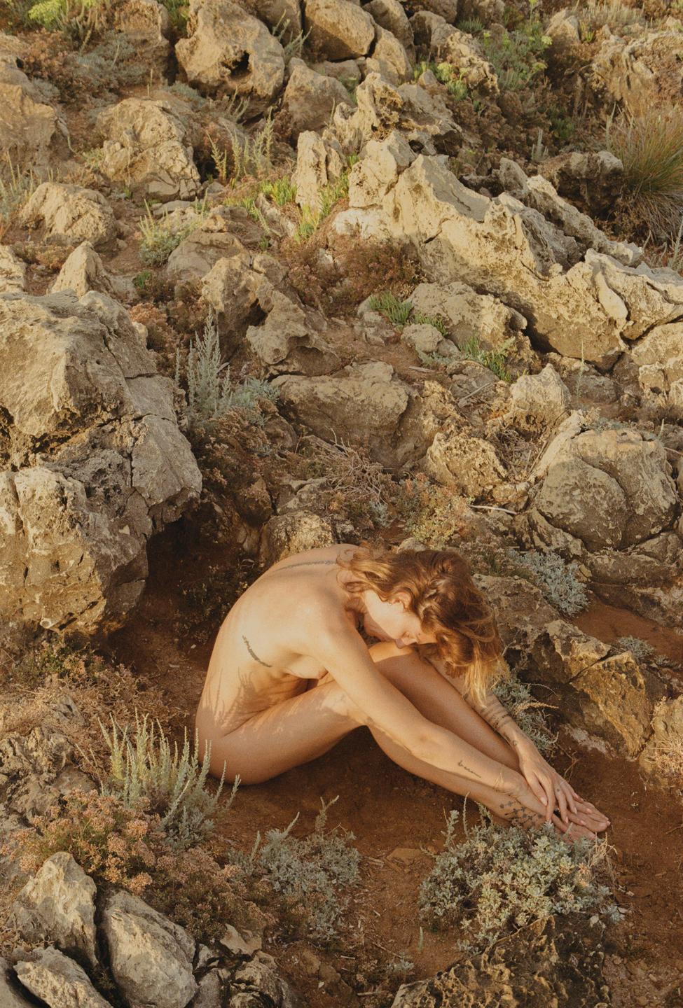 A woman sits curled up on a rocky, arid landscape with sparse vegetation.