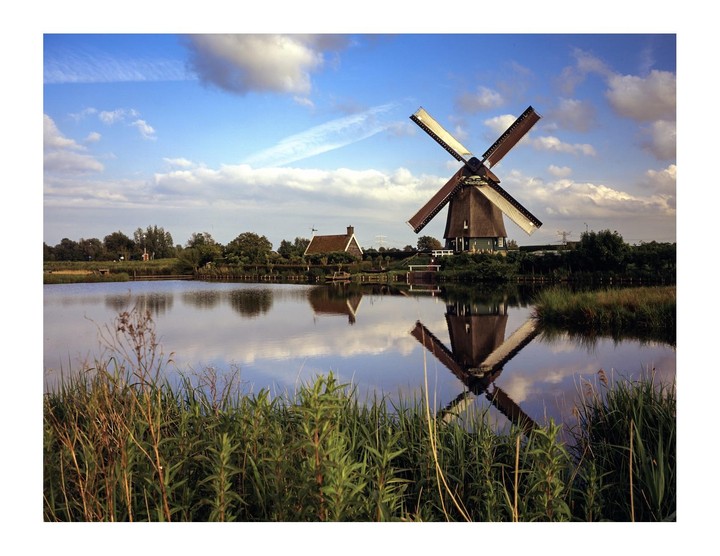 A windmill with a thatched roof stands beside a calm body of water, reflecting its image under a blue sky with scattered clouds.