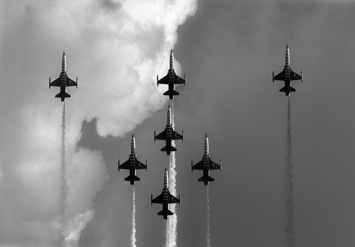 Six jet aircraft fly in formation, leaving smoke trails against a cloudy sky.