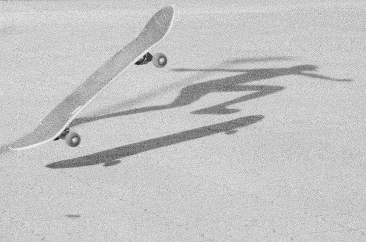 A skateboard is captured mid-air with its shadow and the shadow of a person on a textured surface.