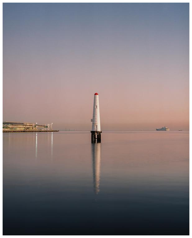 A white lighthouse with a red top stands in calm water with a building and a ship in the distance under a pastel sky.