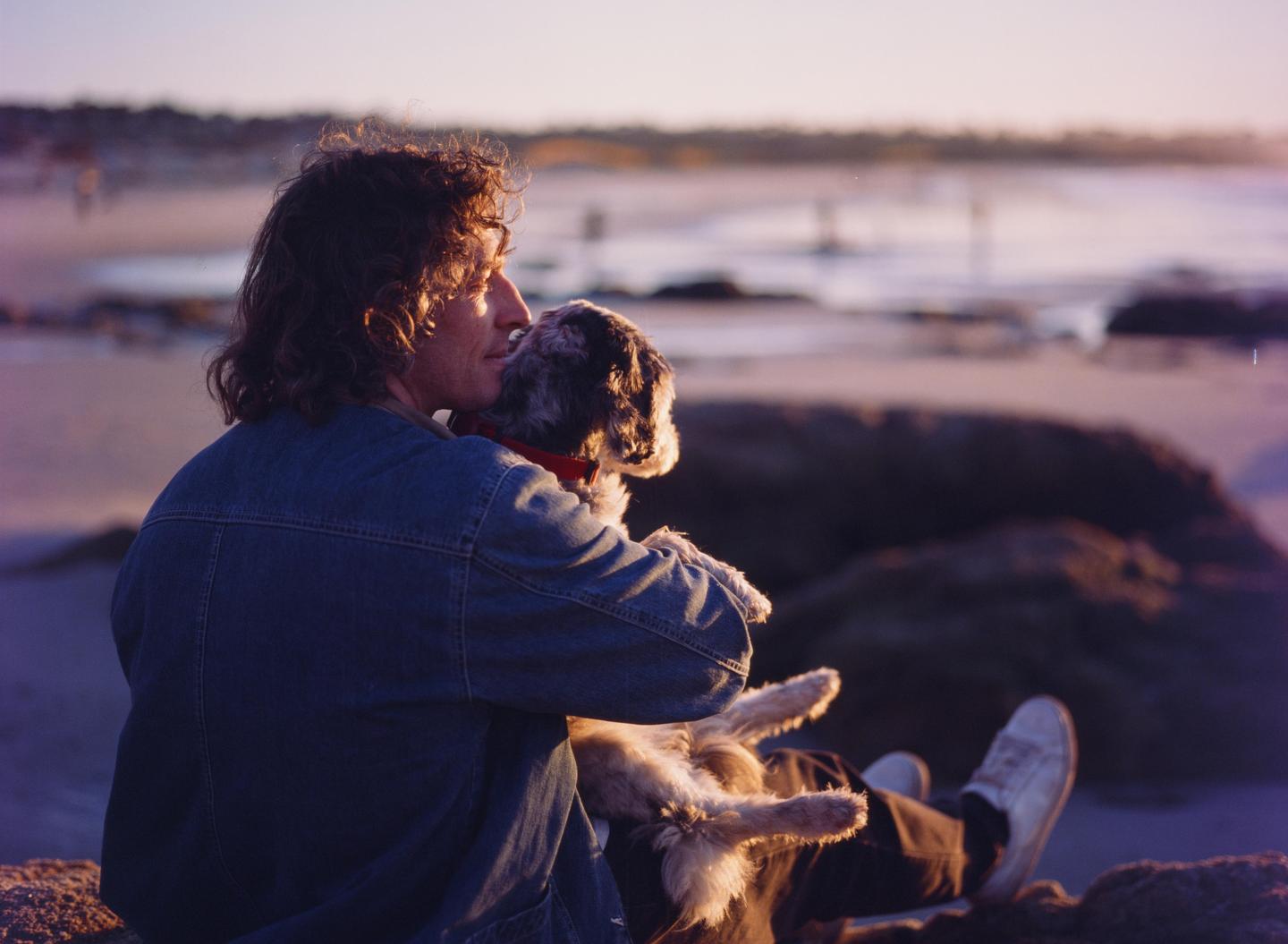 A man sits on a beach at sunset, holding and looking at his dog.