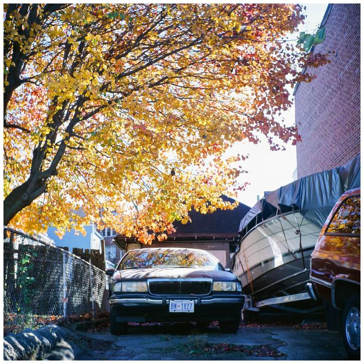 A car is parked under a tree with yellow autumn foliage next to a boat covered with a tarp.