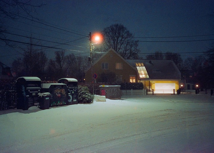 A snow-covered street with a house and recycling bins is illuminated by a street light at night.
