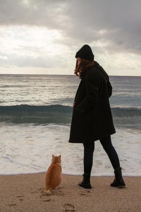 A woman and a cat stand on a sandy beach looking out at the ocean under a cloudy sky.