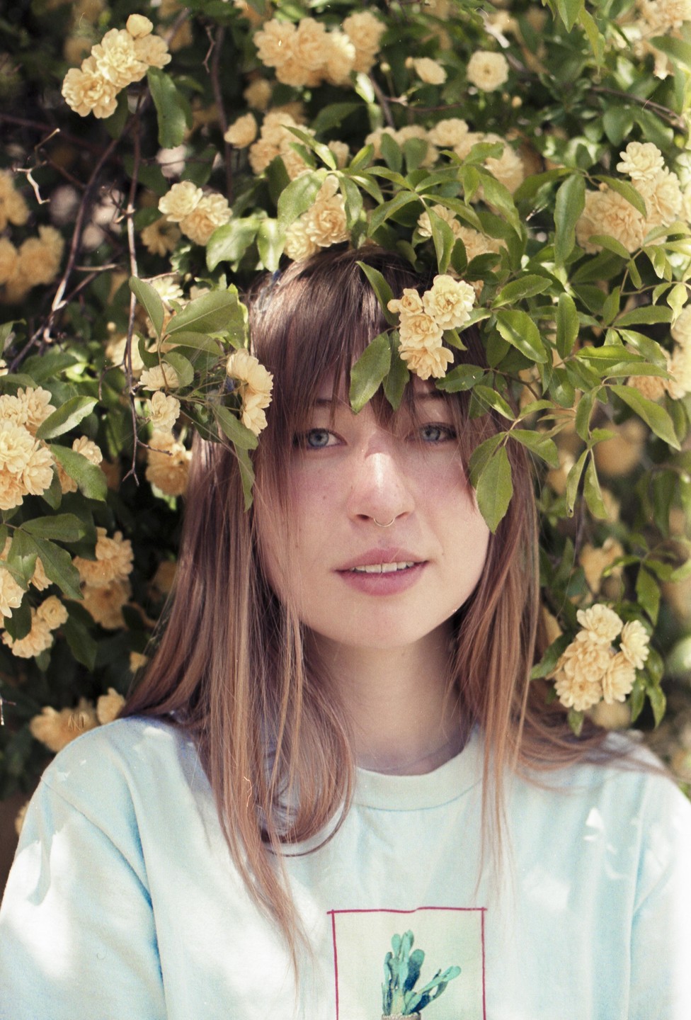 A woman with long hair and bangs looks at the viewer, surrounded by yellow flowers and green leaves.