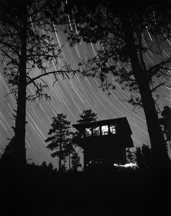 A tall building with illuminated windows stands silhouetted against a night sky filled with star trails and surrounded by trees.