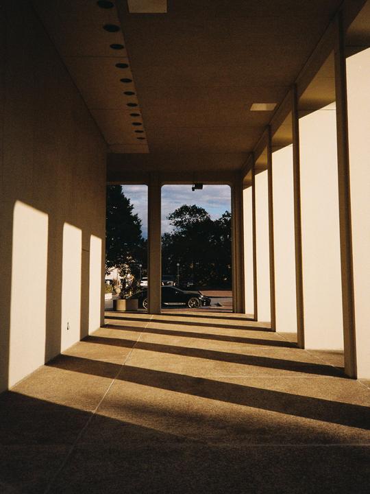 A long hallway with columns and shadows leads to an outdoor area with a car and trees.