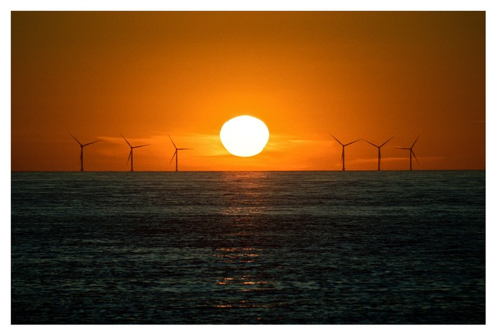 The sun sets over the ocean with several wind turbines visible on the horizon.
