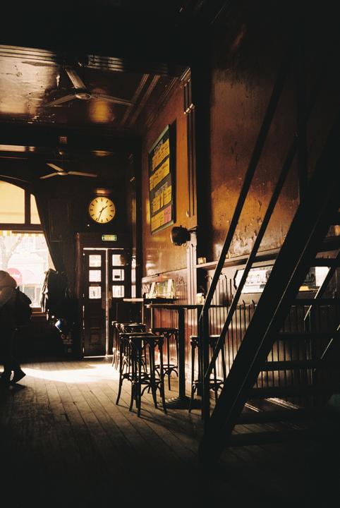 Sunlight streams into a dark, wood-paneled bar with high tables and stools, a clock on the wall, and a staircase on the right.