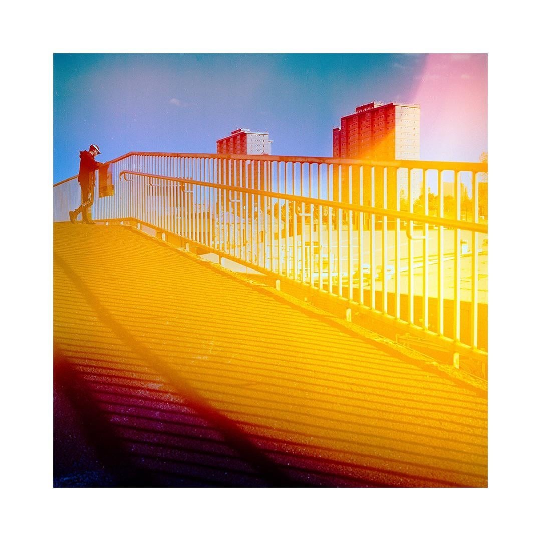 A person stands on a bridge with a railing, looking out at distant buildings under a bright sky.