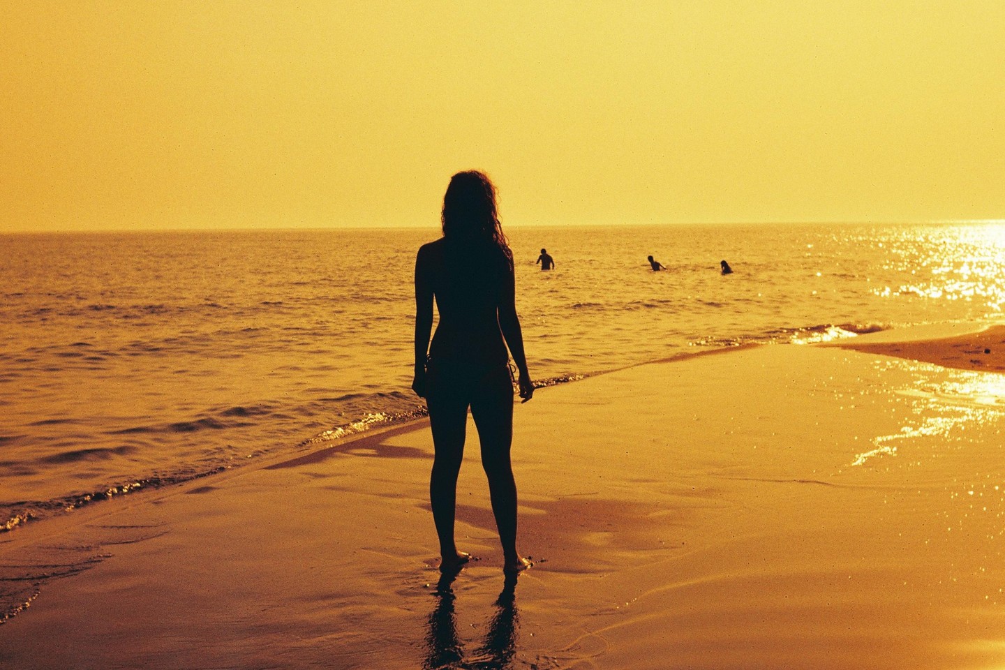 A woman stands on a beach looking out at the ocean with other people swimming in the distance during sunset.
