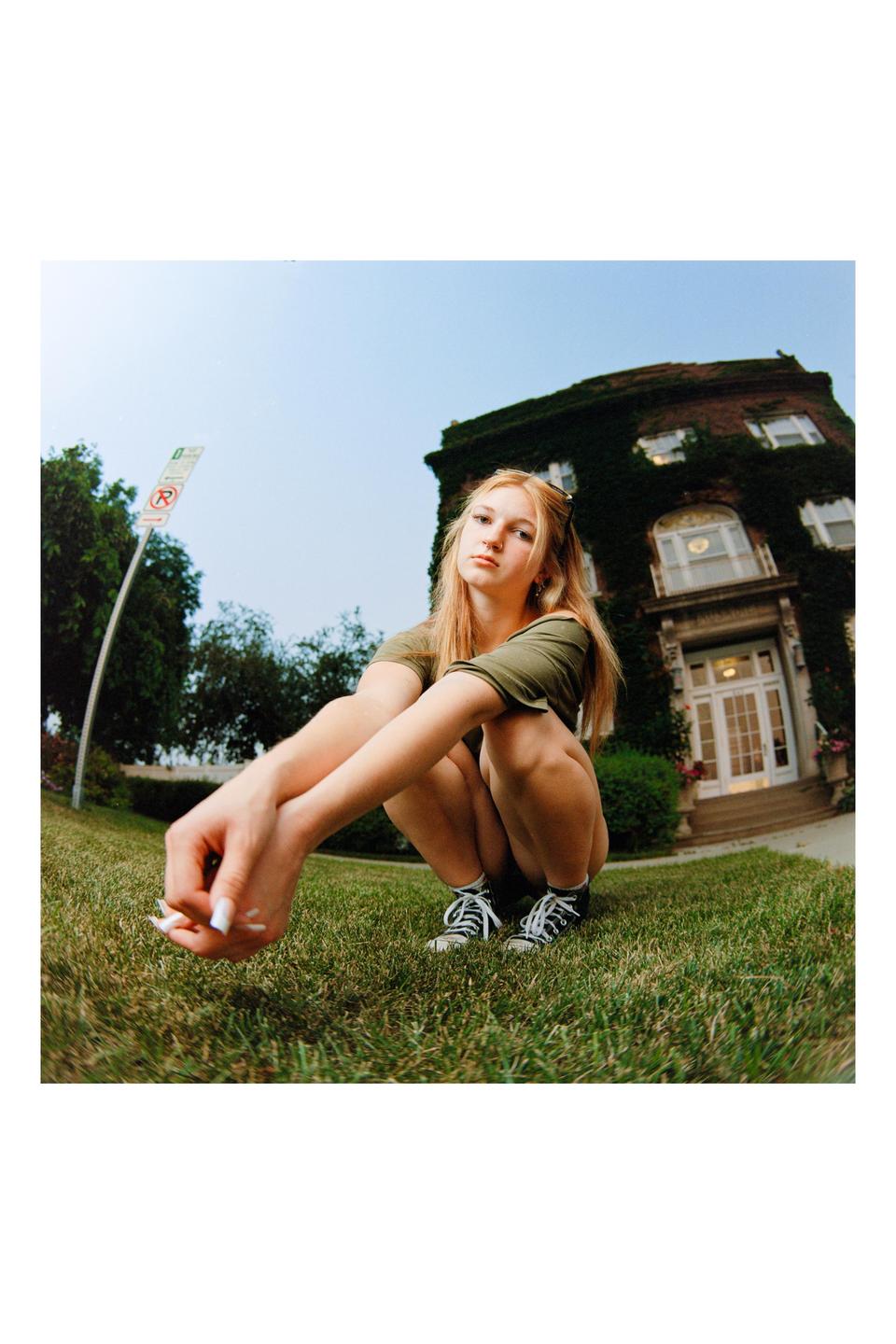 A young woman squats on a grassy lawn in front of an ivy-covered building.