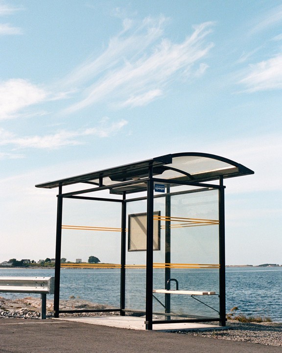 A bus stop with a clear roof and glass walls stands next to a body of water under a blue sky with wispy clouds.