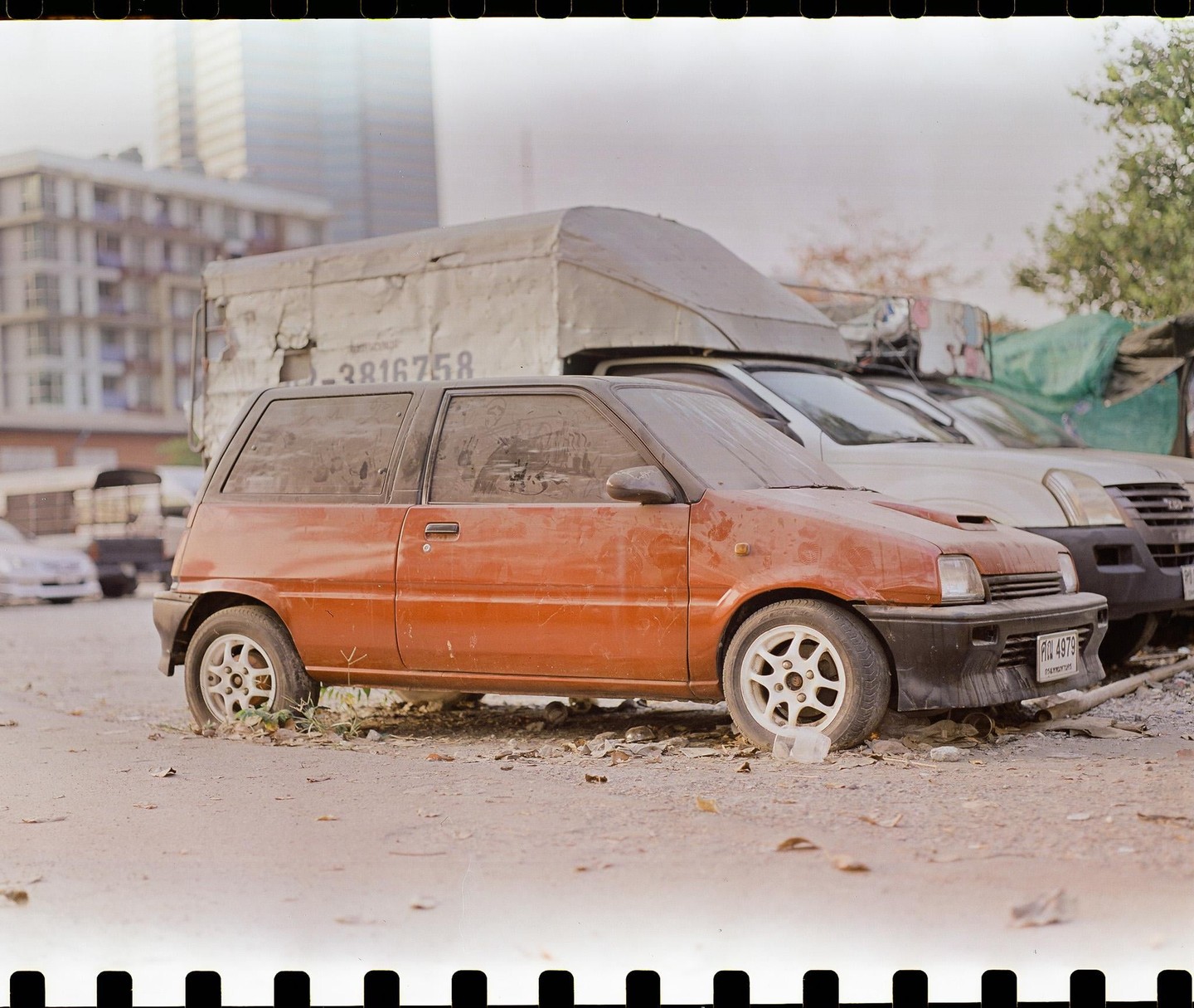 A dusty orange car is parked on a dirt lot with other vehicles and buildings in the background.