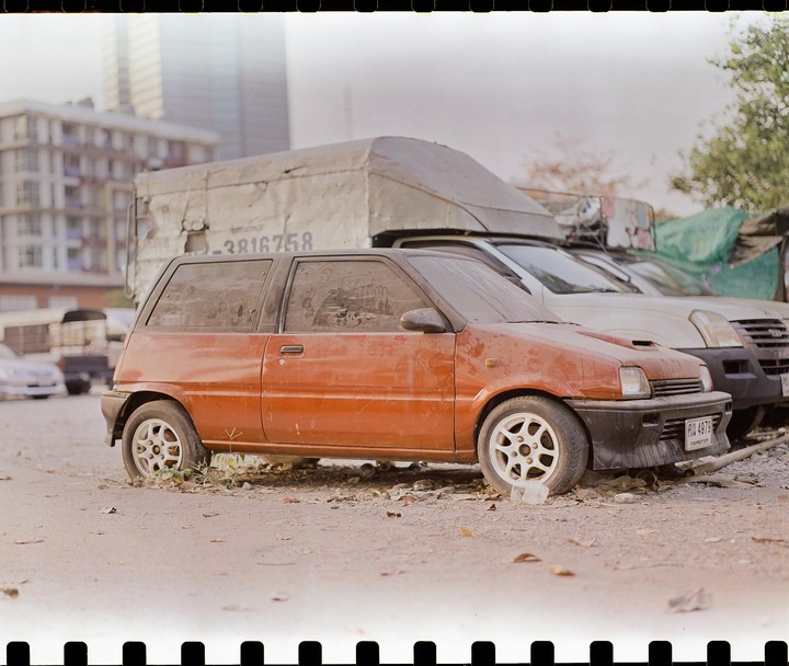A dusty orange car is parked on a dirt lot with other vehicles and buildings in the background.