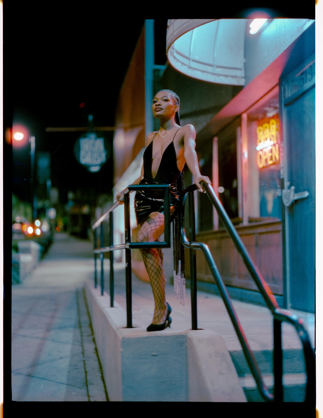 A woman in a black dress and fishnet stockings stands on a ledge next to a railing at night.
