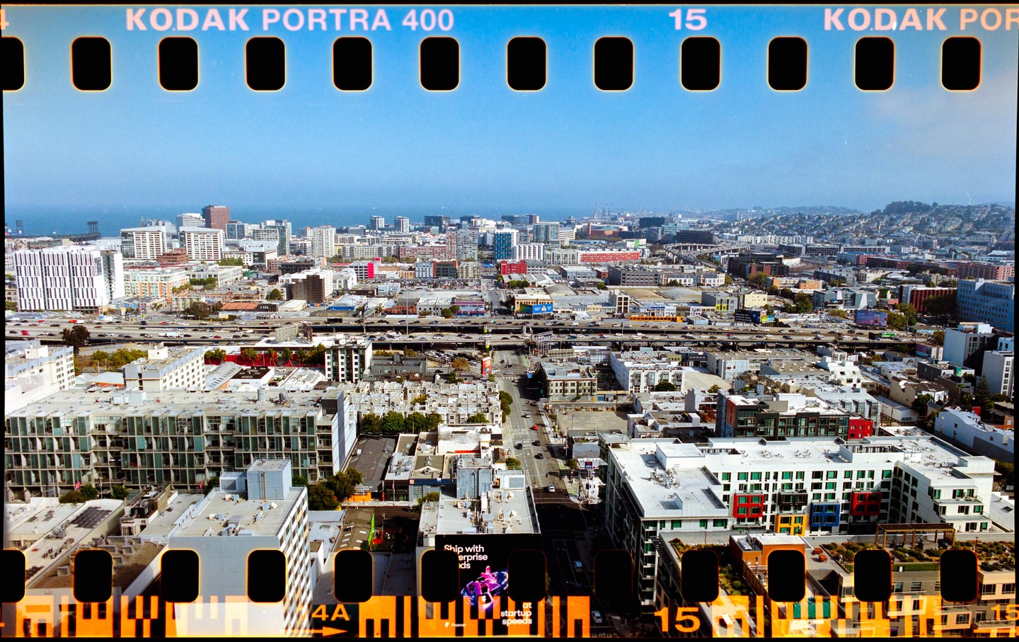 An aerial view shows a dense urban landscape with a highway cutting through buildings and a body of water in the distance under a clear sky.