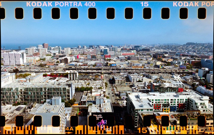 An aerial view shows a dense urban landscape with a highway cutting through buildings and a body of water in the distance under a clear sky.