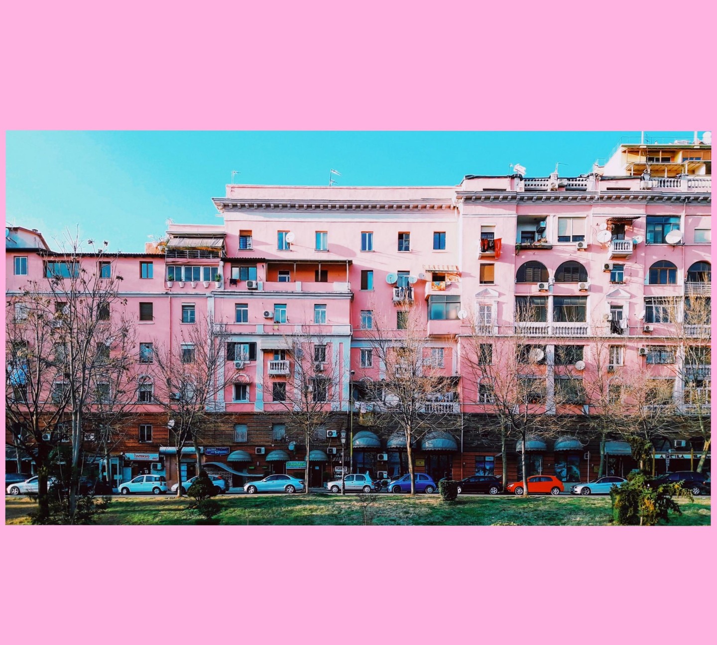 A row of pink buildings with bare trees and parked cars in front of them is visible under a clear blue sky.