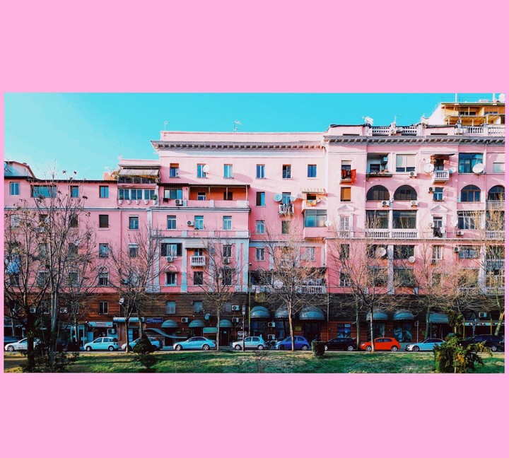 A row of pink buildings with bare trees and parked cars in front of them is visible under a clear blue sky.