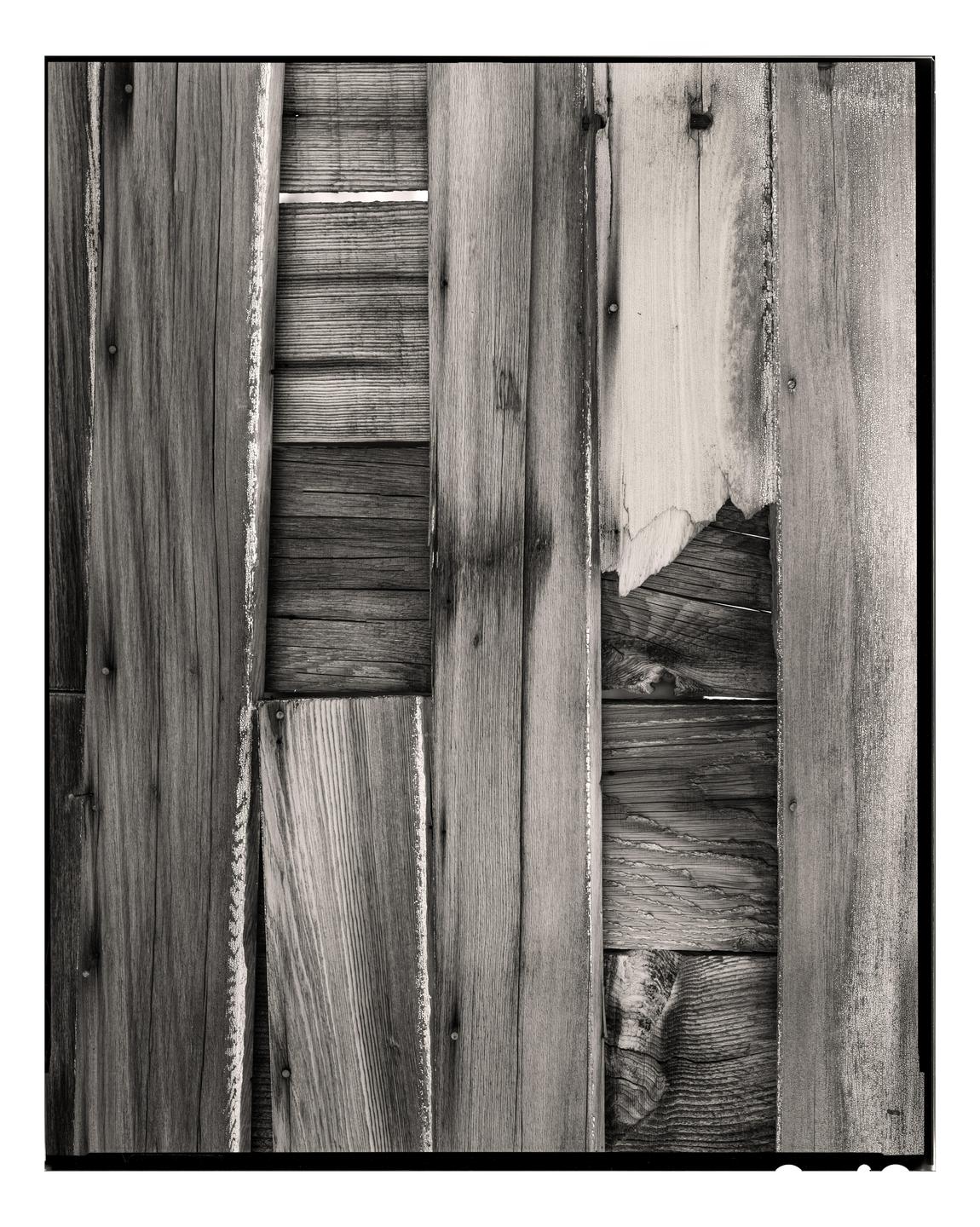 A black and white close-up shows a weathered wooden fence with several planks missing or broken, revealing more wood behind.