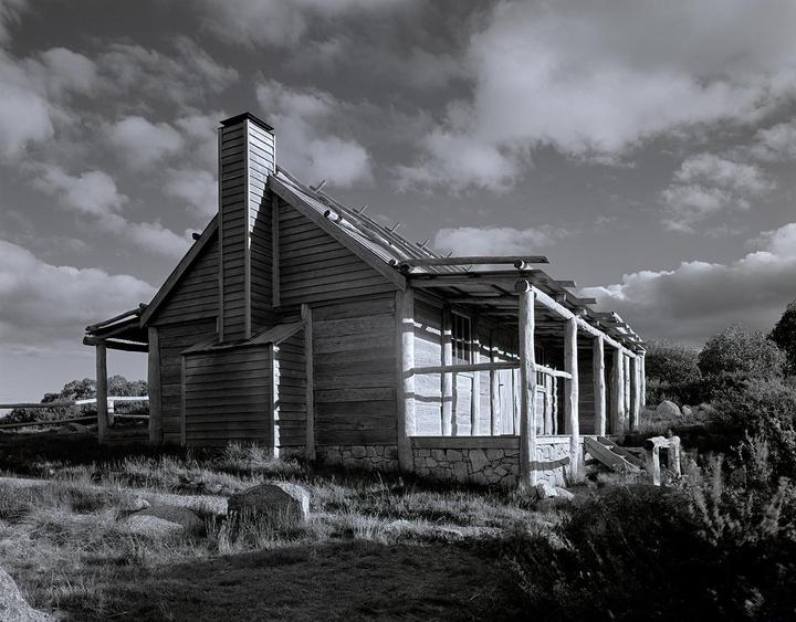 A wooden hut with a stone foundation and a porch stands on a grassy hill under a cloudy sky.