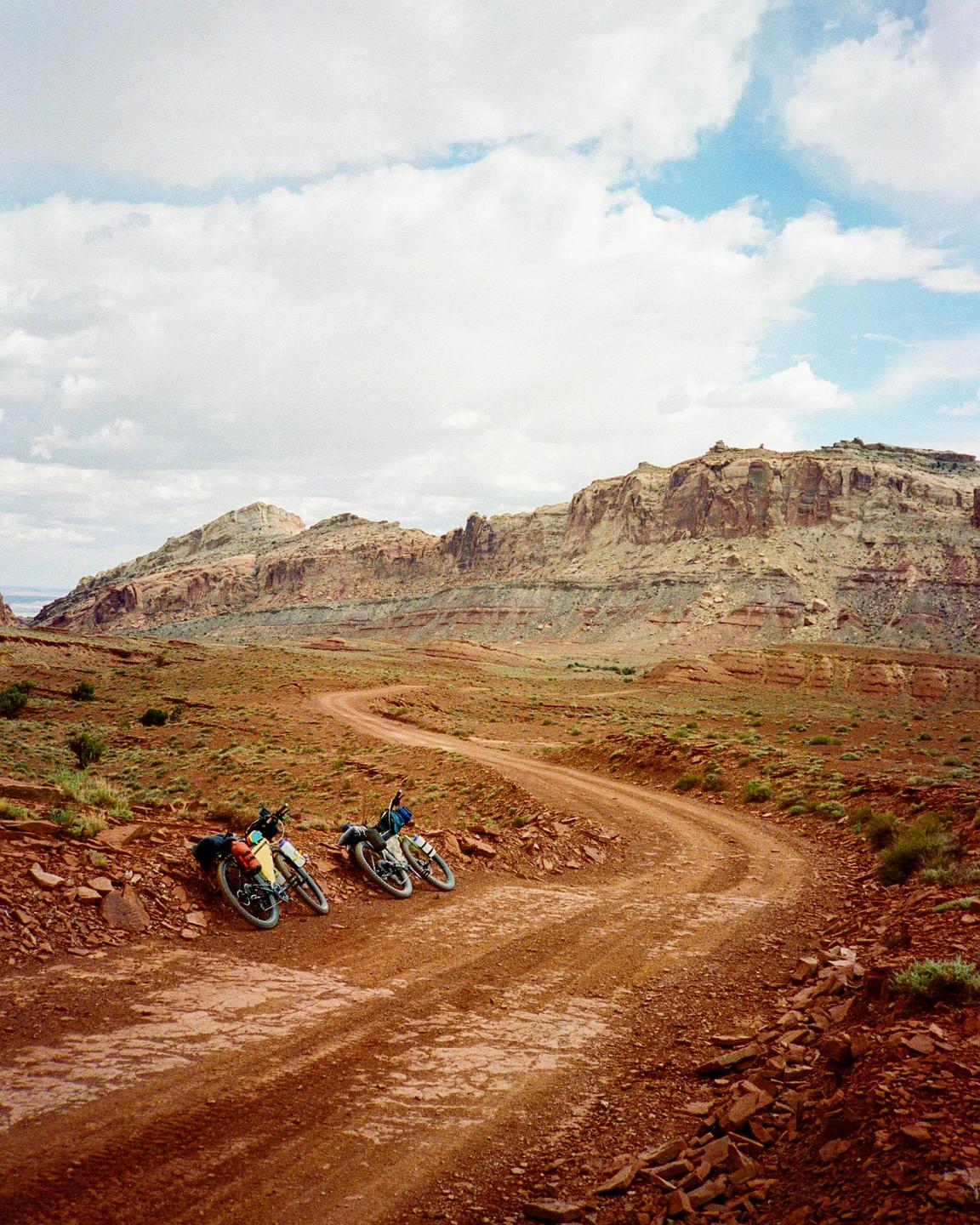 Two bicycles are parked on the side of a winding dirt road in a desert landscape with mountains in the background.